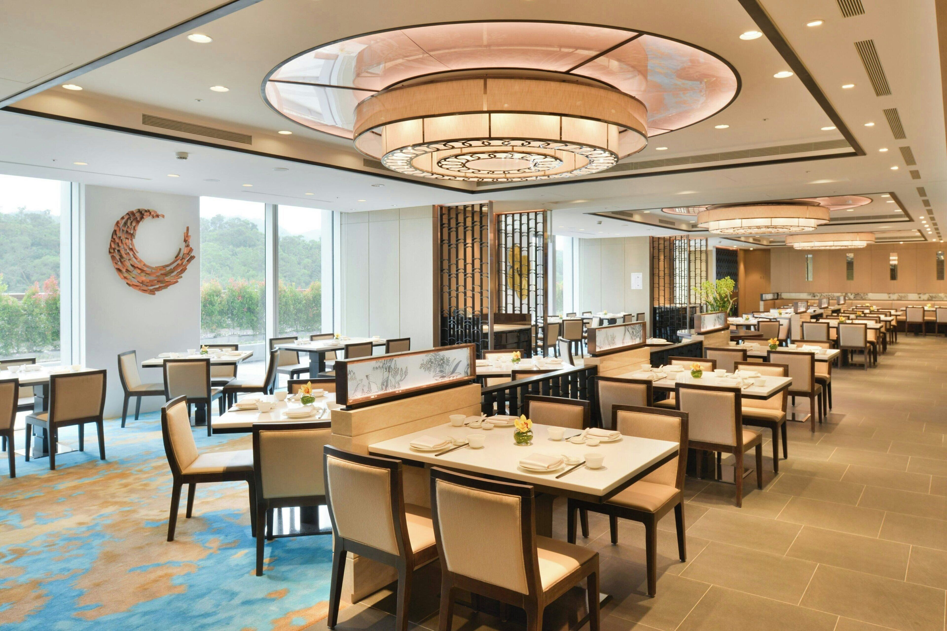 A spacious dining area with multiple tables and chairs, large windows offering a view of greenery, and a modern ceiling design.