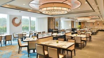 A spacious dining area with multiple tables and chairs, large windows offering a view of greenery, and a modern ceiling design.