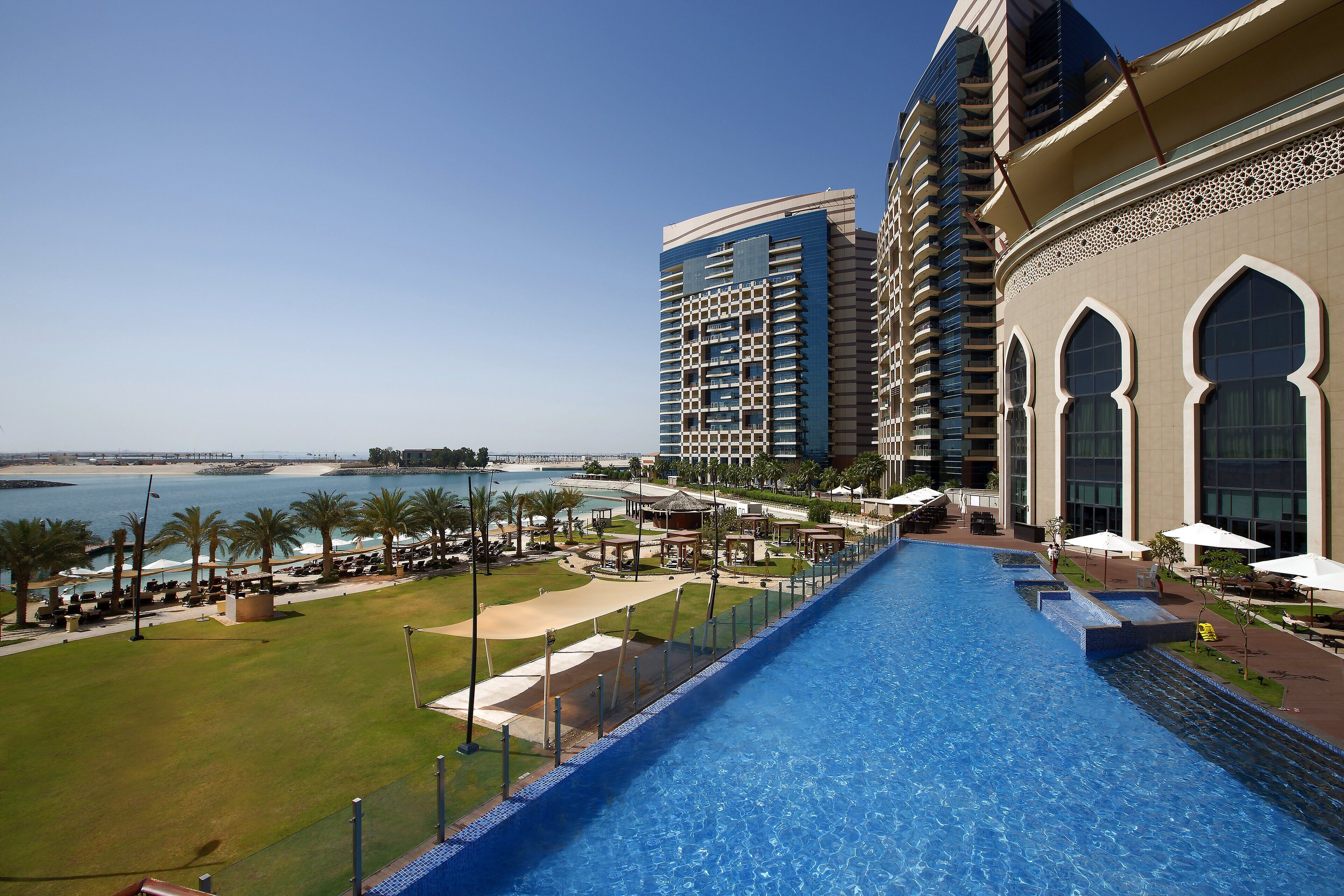 A swimming pool with a view of the sea, surrounded by palm trees and a building with arched windows.