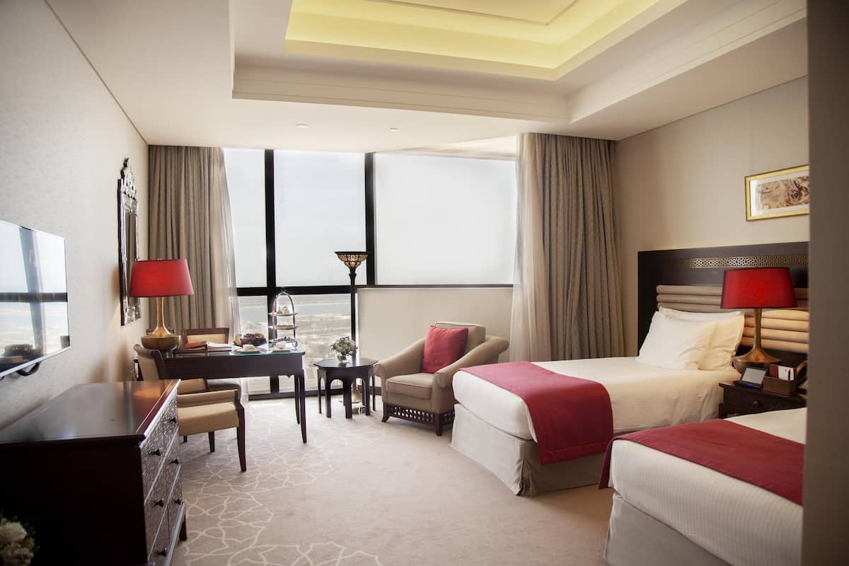 A hotel room with two beds, a desk, a chair, a TV, and a view of the city.