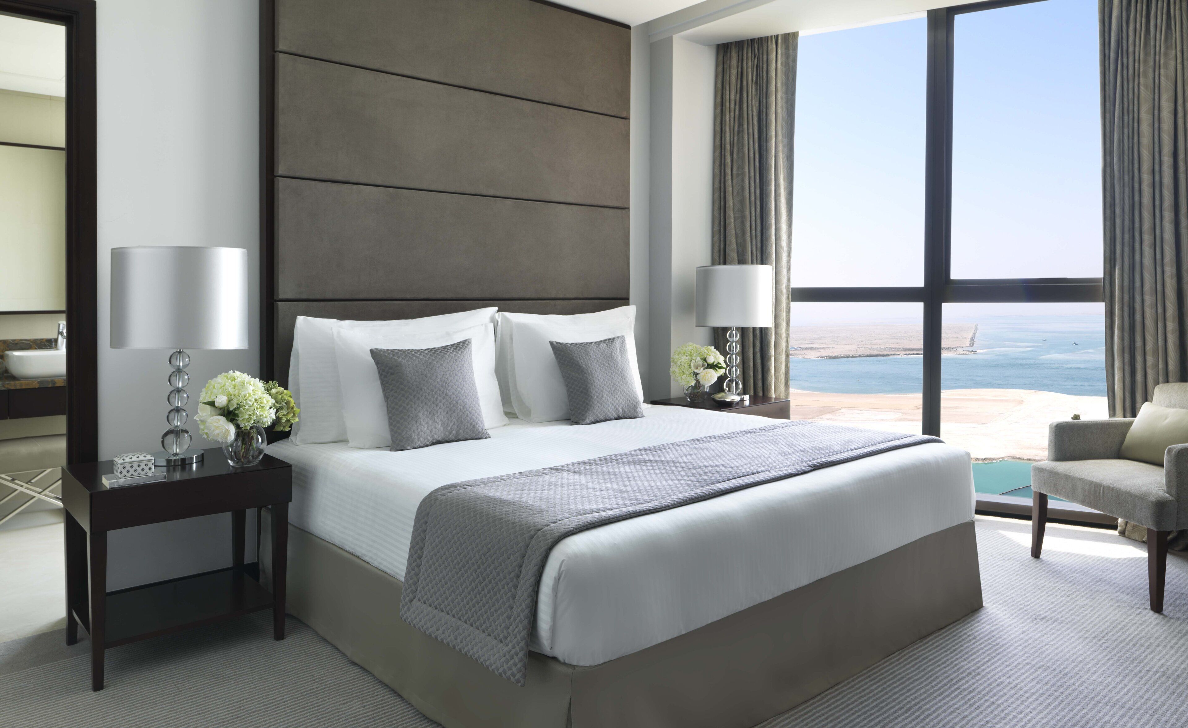 A modern hotel room with a large bed, two bedside tables, a nightstand, a view of the sea, and a balcony.