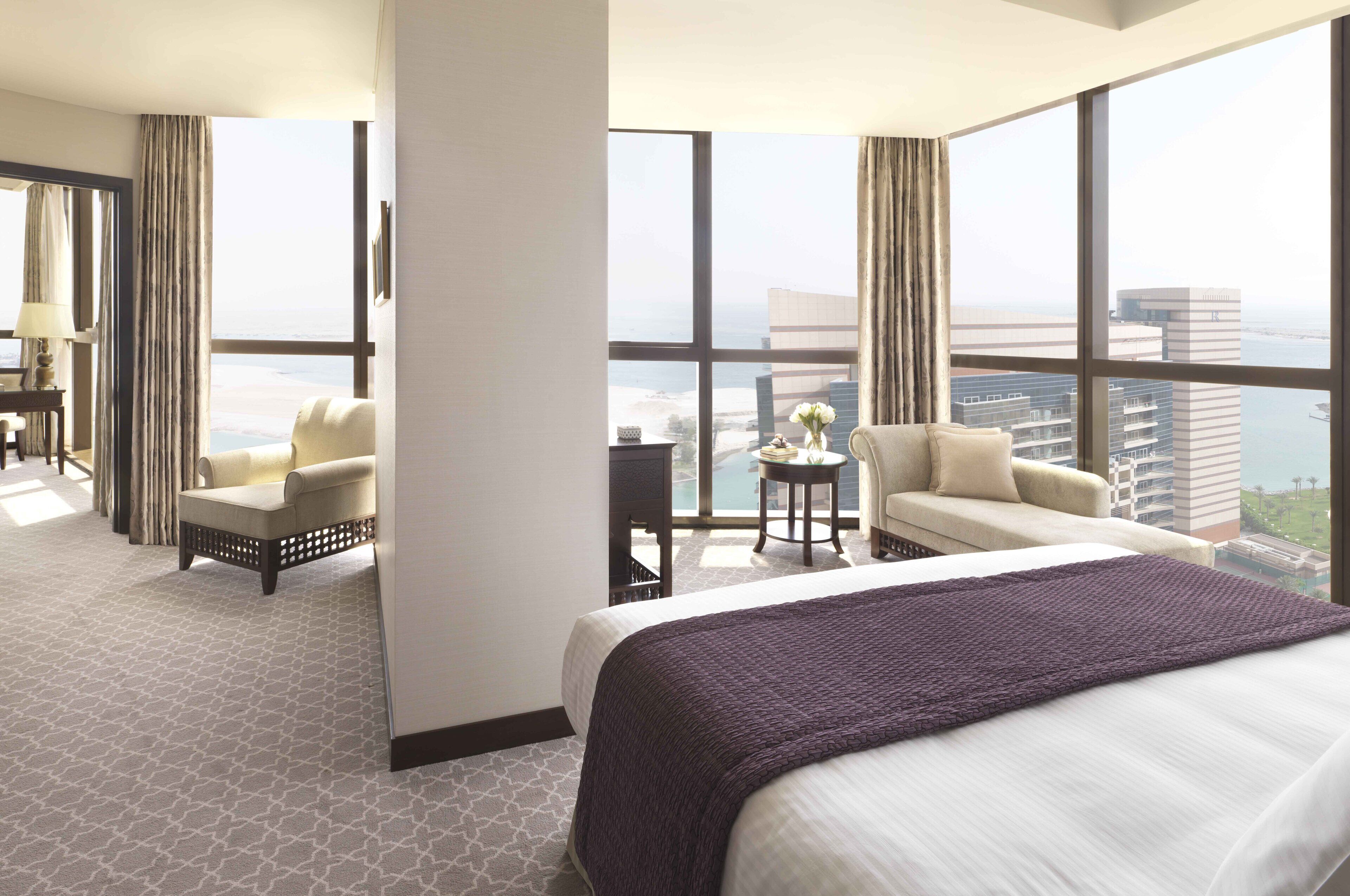 Habitación de hotel con una cama grande, dos sillones, una mesita con un florero y vistas a la ciudad y al agua.