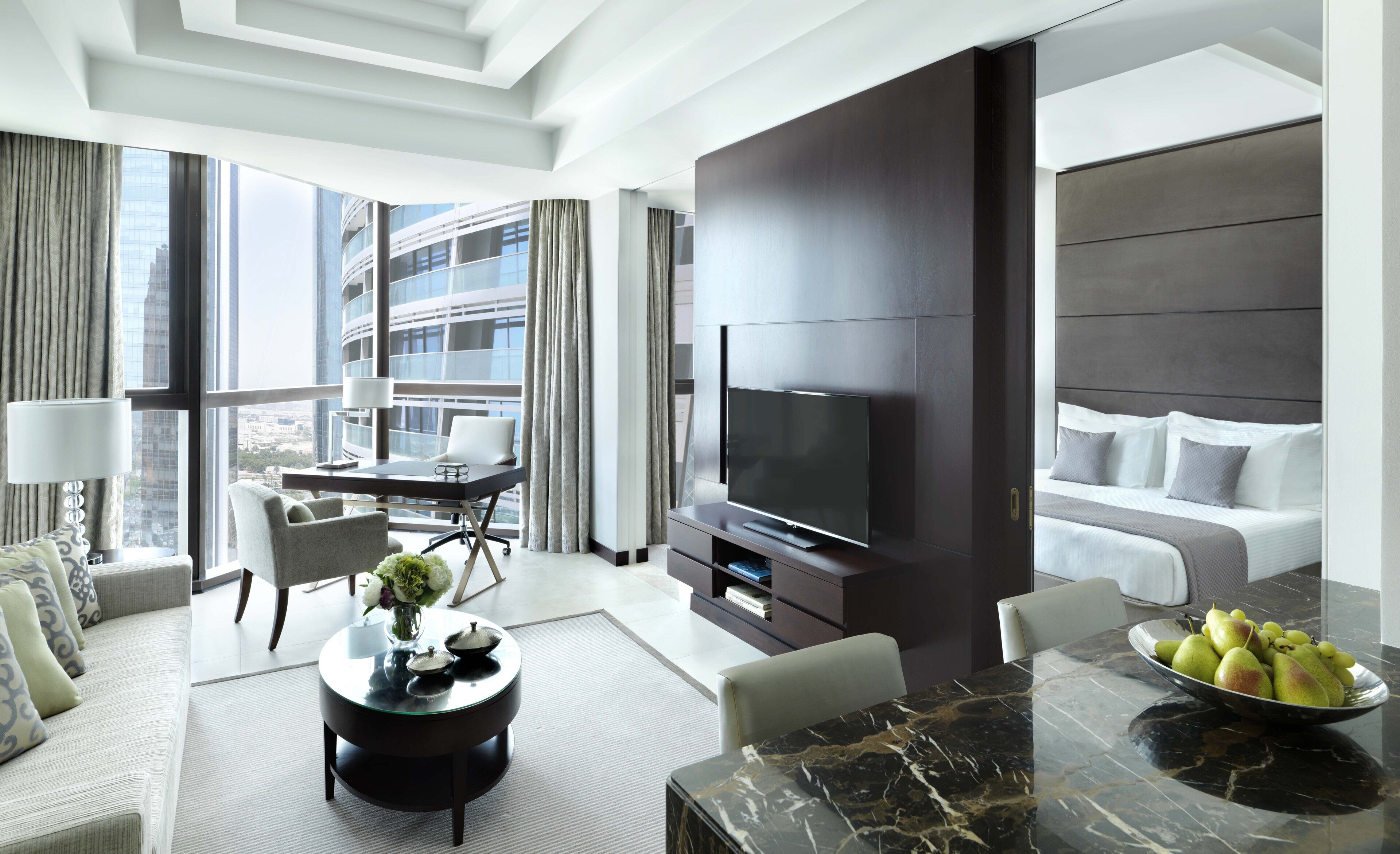 A modern hotel room with a large TV, a dining area, a sofa, and a balcony with a city view.