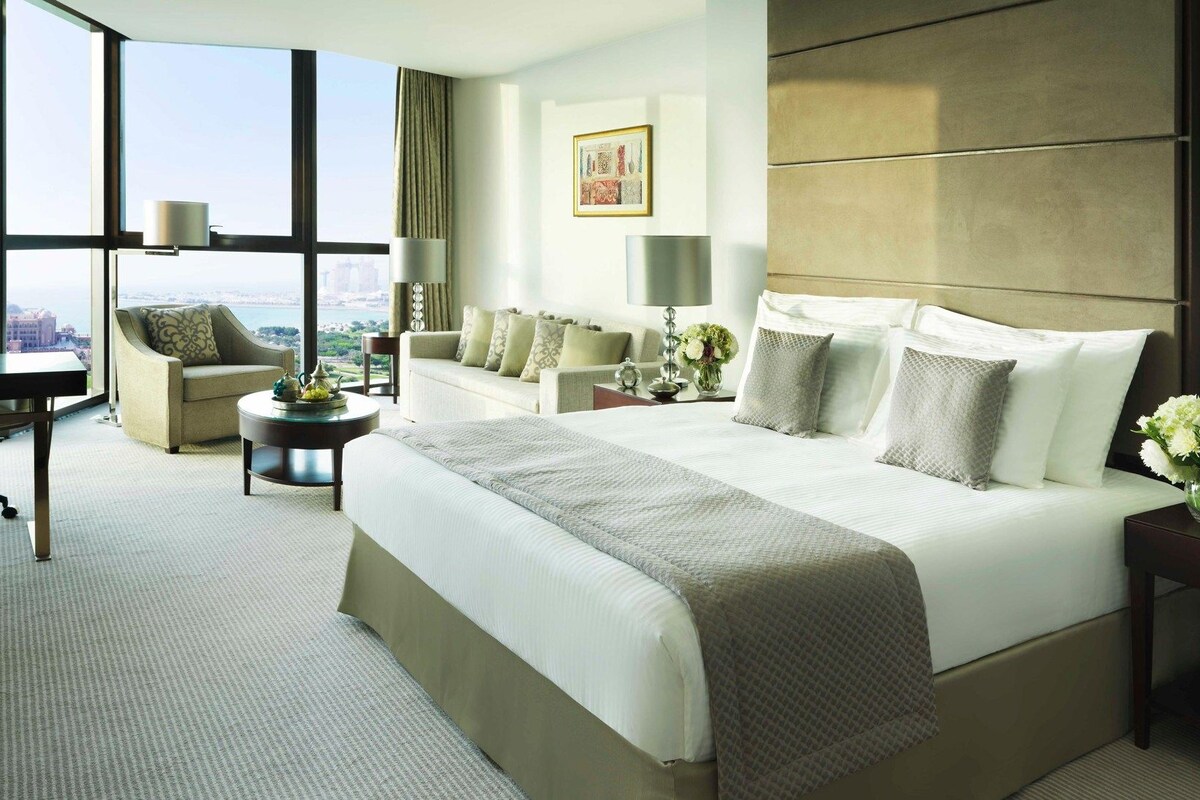 Une chambre d’hôtel avec un grand lit, deux fauteuils, un bureau et une vue sur la ville.