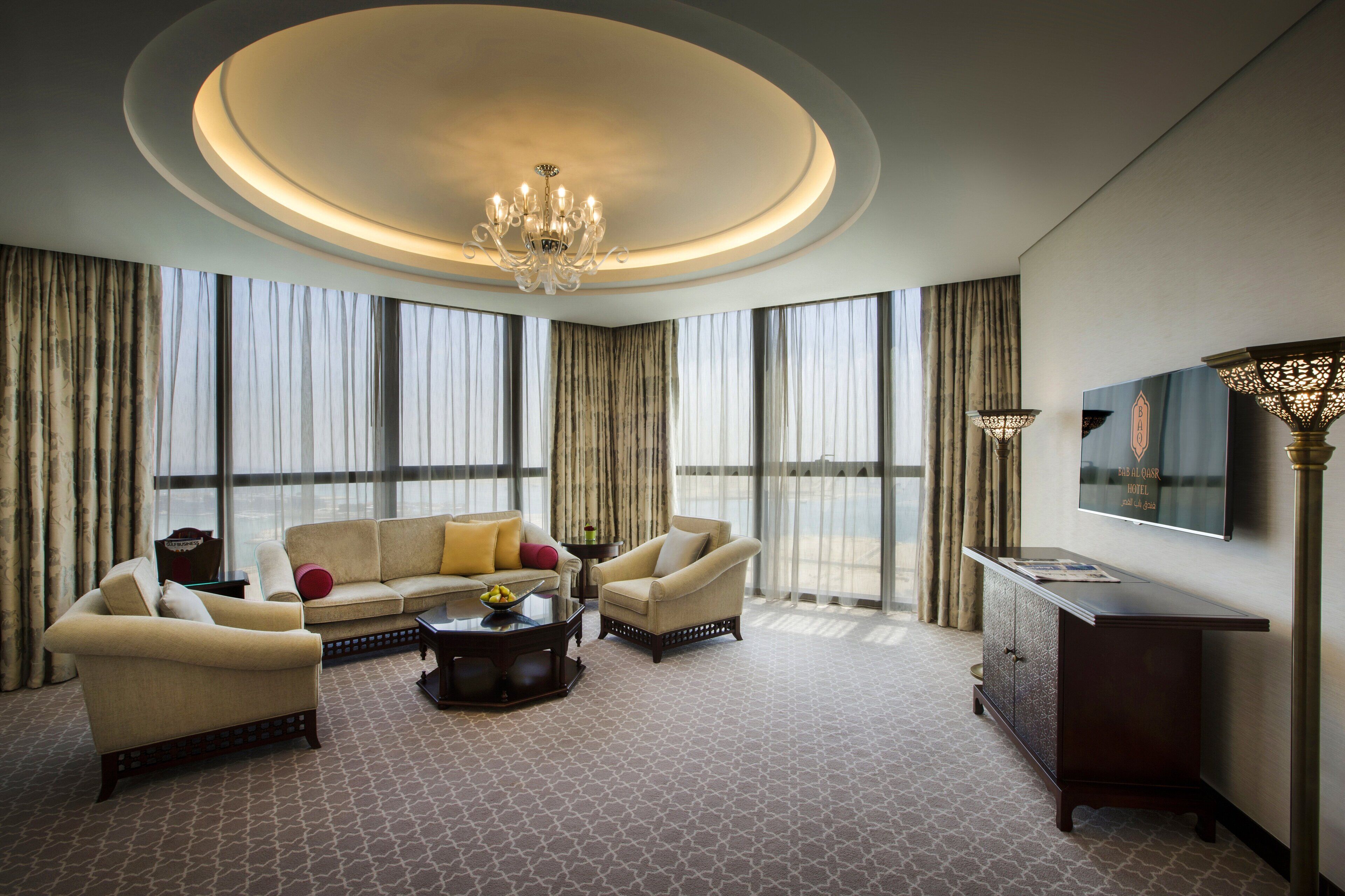 A spacious living room with a large chandelier, a sofa set, a coffee table, and a TV mounted on the wall.
