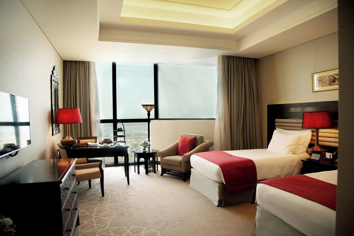 Habitación de hotel con cama, escritorio, silla, televisor y ventanas amplias.