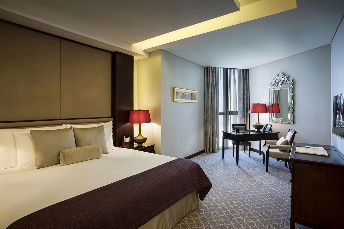 Une chambre d’hôtel avec un grand lit, un bureau, deux chaises, une lampe de bureau, un miroir et un tableau encadré au mur.
