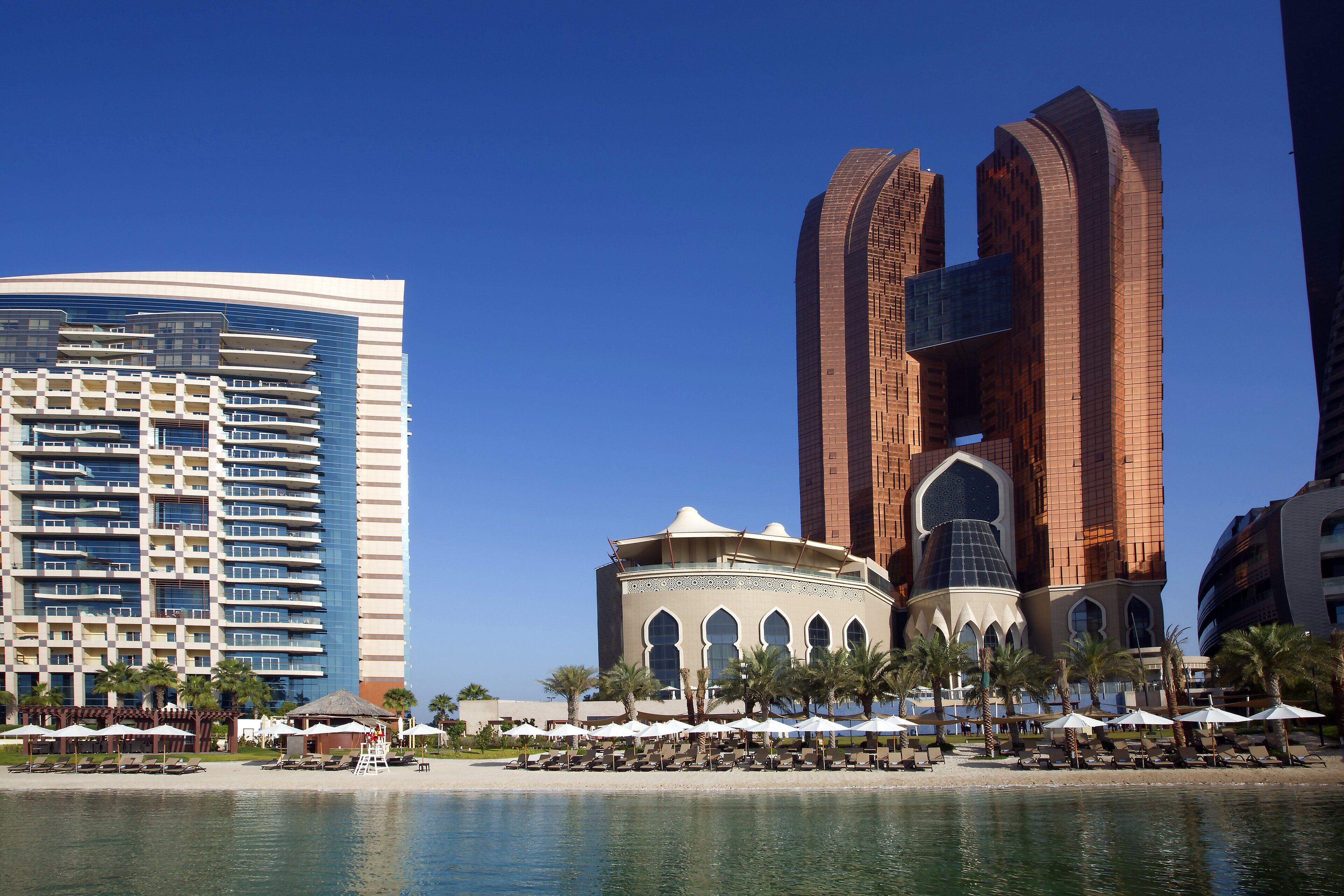 Un hotel frente a la playa con un edificio moderno, una mezquita con cúpula y palmeras.