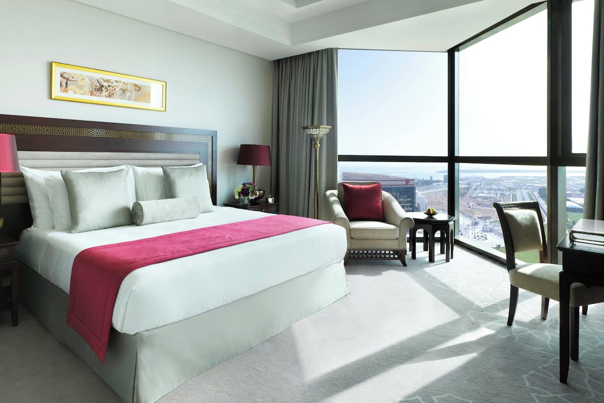 A hotel room with a large bed, two bedside tables, a desk, a chair, a view of the city, and a large window.
