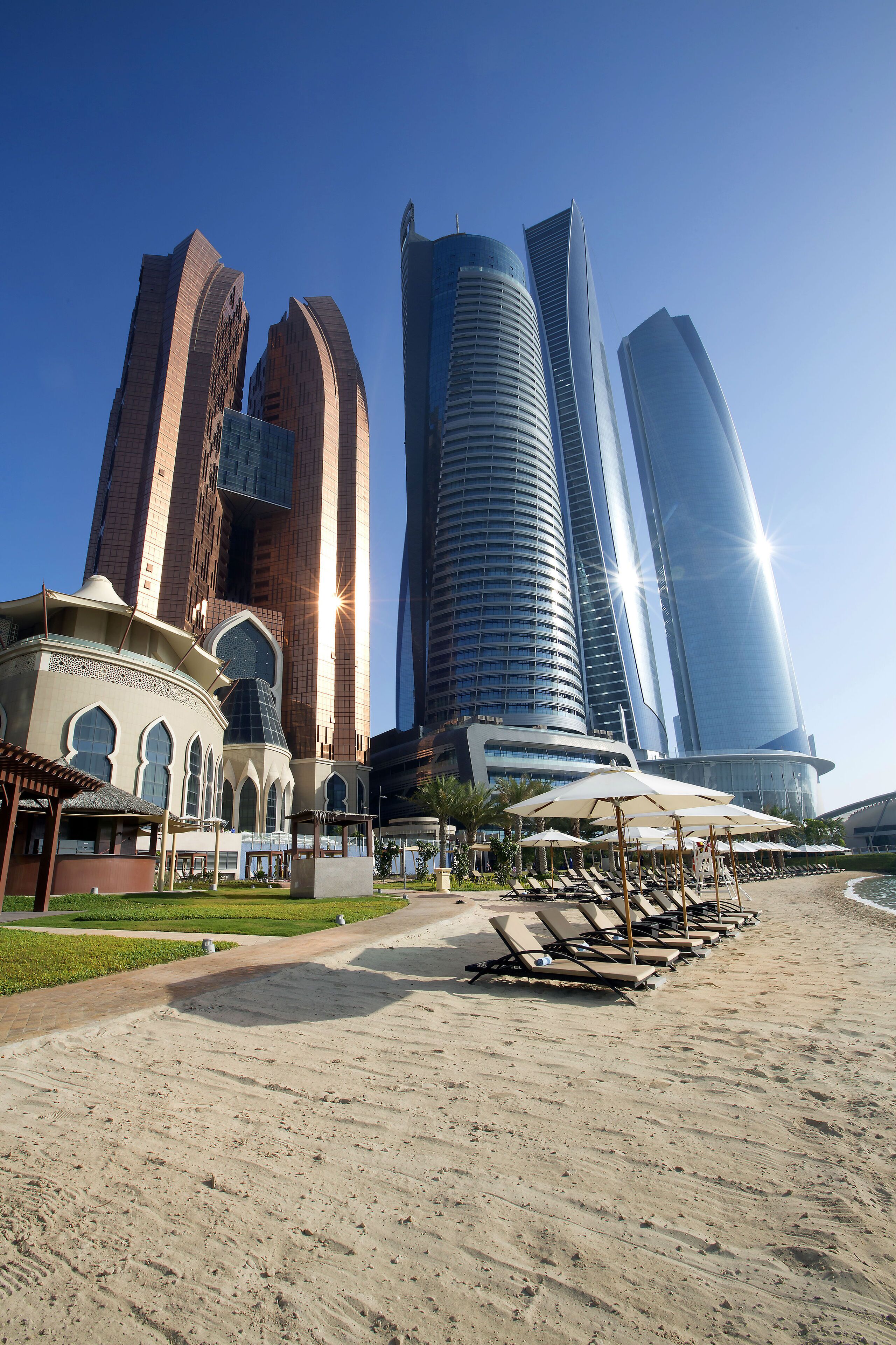A modern hotel with a prominent curved glass facade, a traditional building with arched windows, and a sandy beach area with sun loungers.