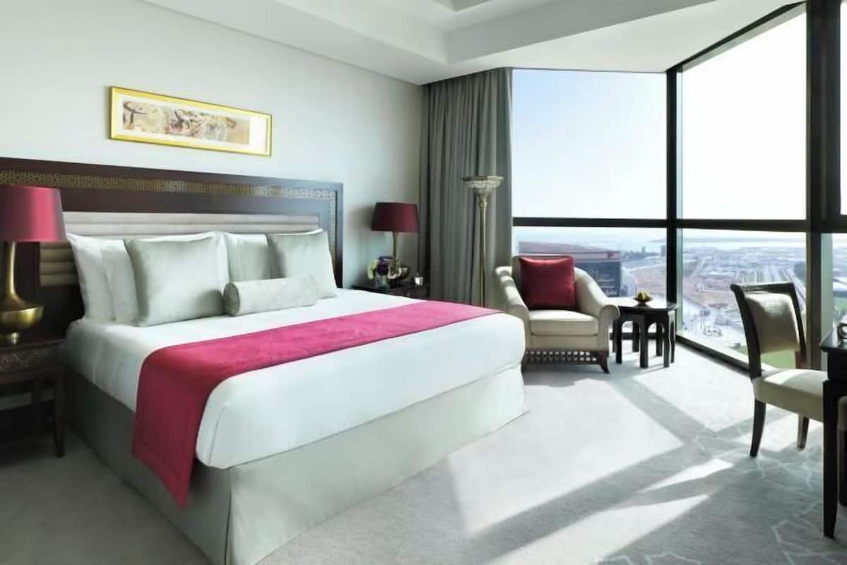 A hotel room with a large bed, a desk, a chair, a lamp, and a view of the city.