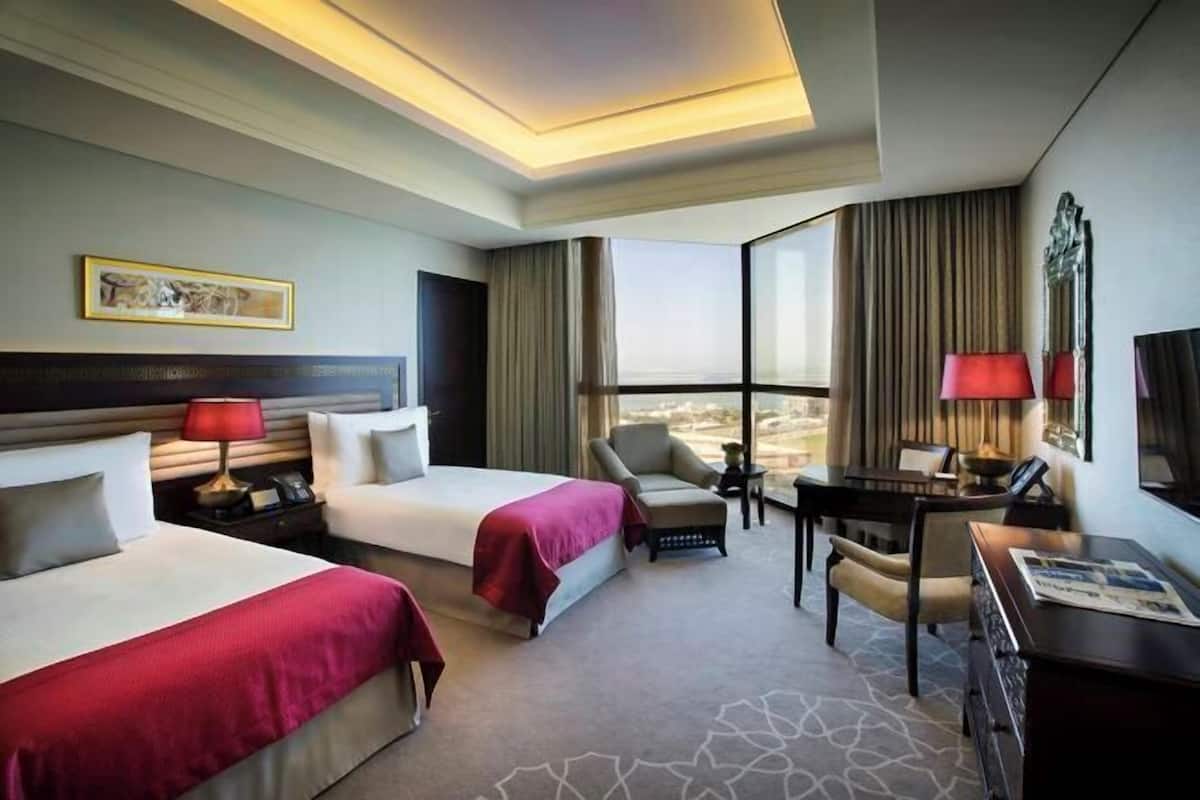 A hotel room with two beds, a desk, a chair, a lamp, and a large window.