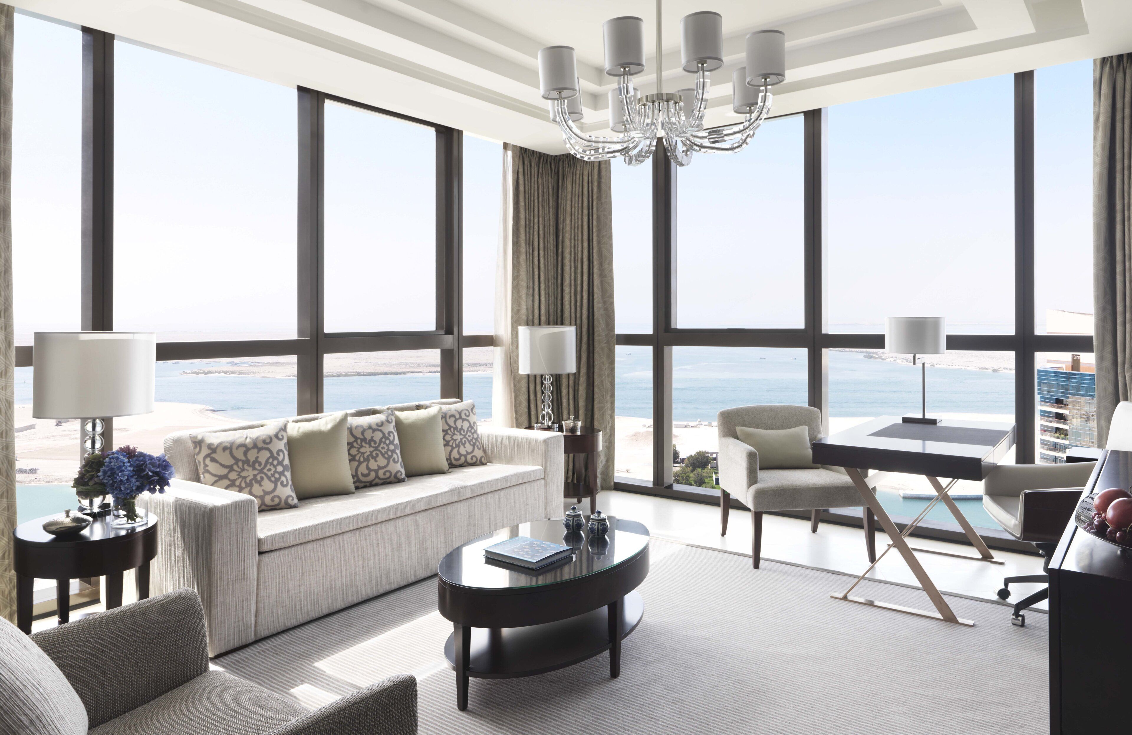 A modern hotel room with a large window overlooking the beach, a sofa, a coffee table, and a desk with a lamp.