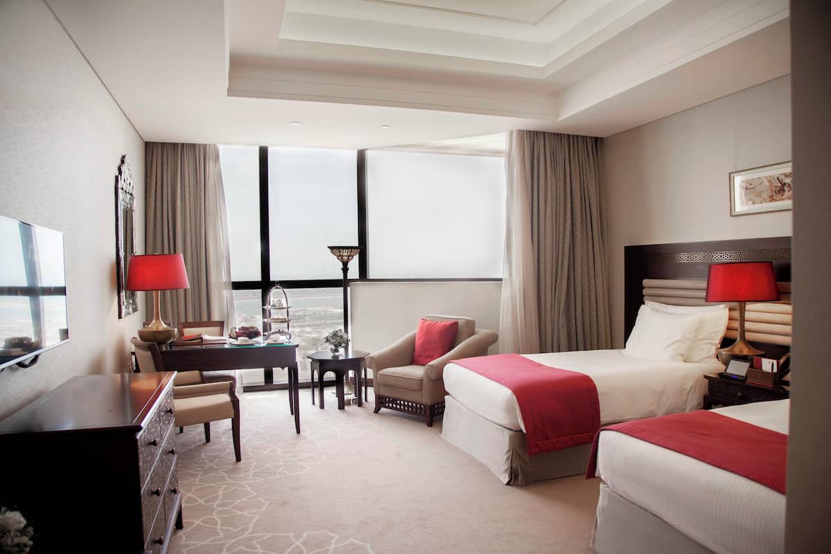 A hotel room with two beds, a desk, a chair, a TV, and a view of the city.
