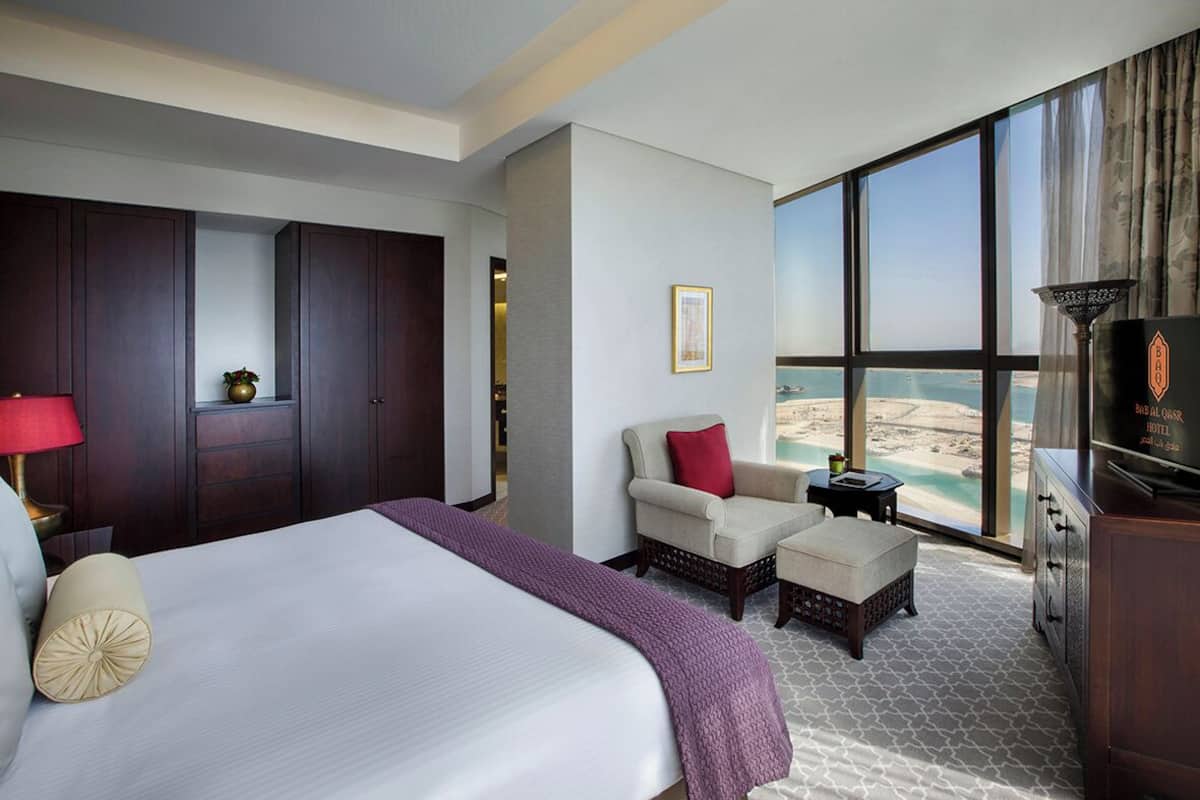A hotel room with a large bed, a seating area with a view of the beach, and a minibar.