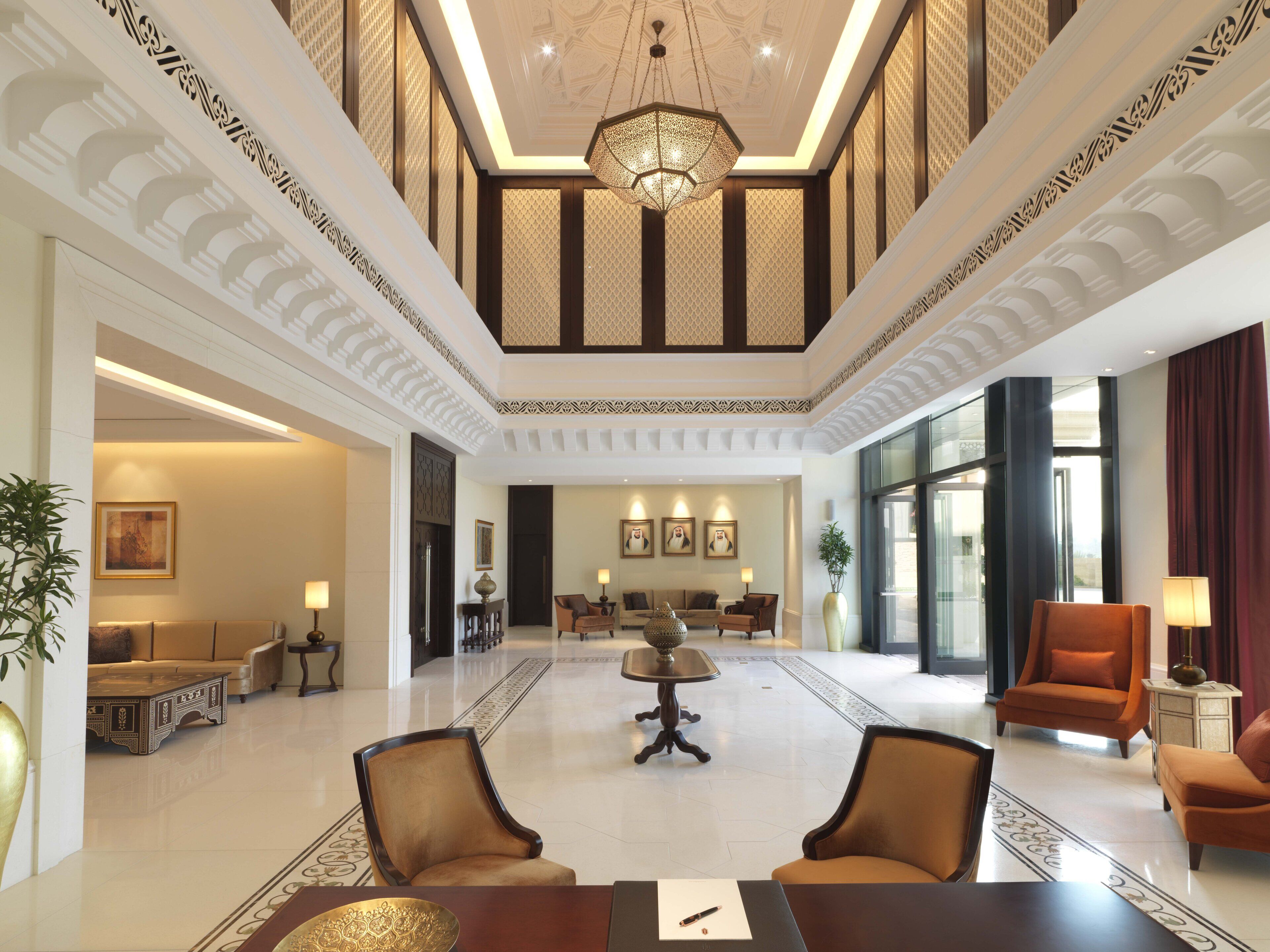 A spacious lobby with a high ceiling, ornate ceiling design, and a chandelier. There are multiple seating areas with sofas and armchairs, a coffee table, and a dining table with chairs.