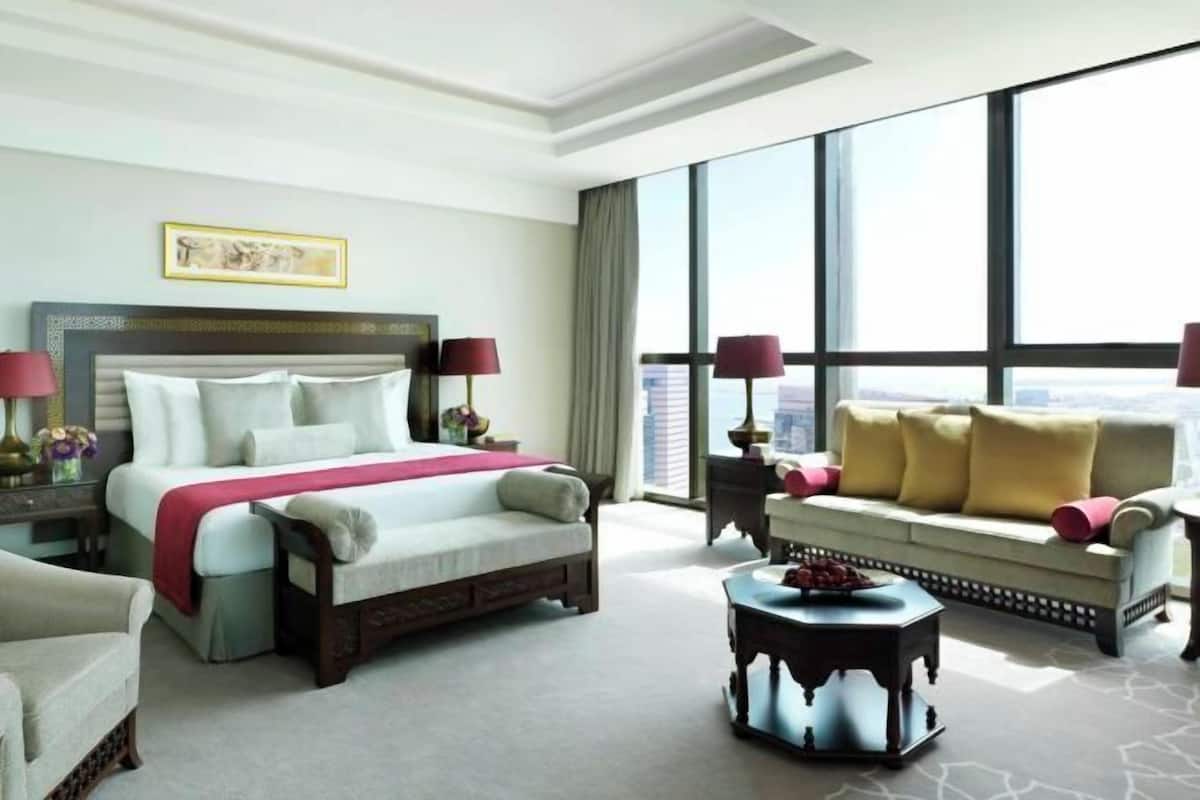 A hotel room with a large bed, a sofa, a small table, and a large window offering a city view.