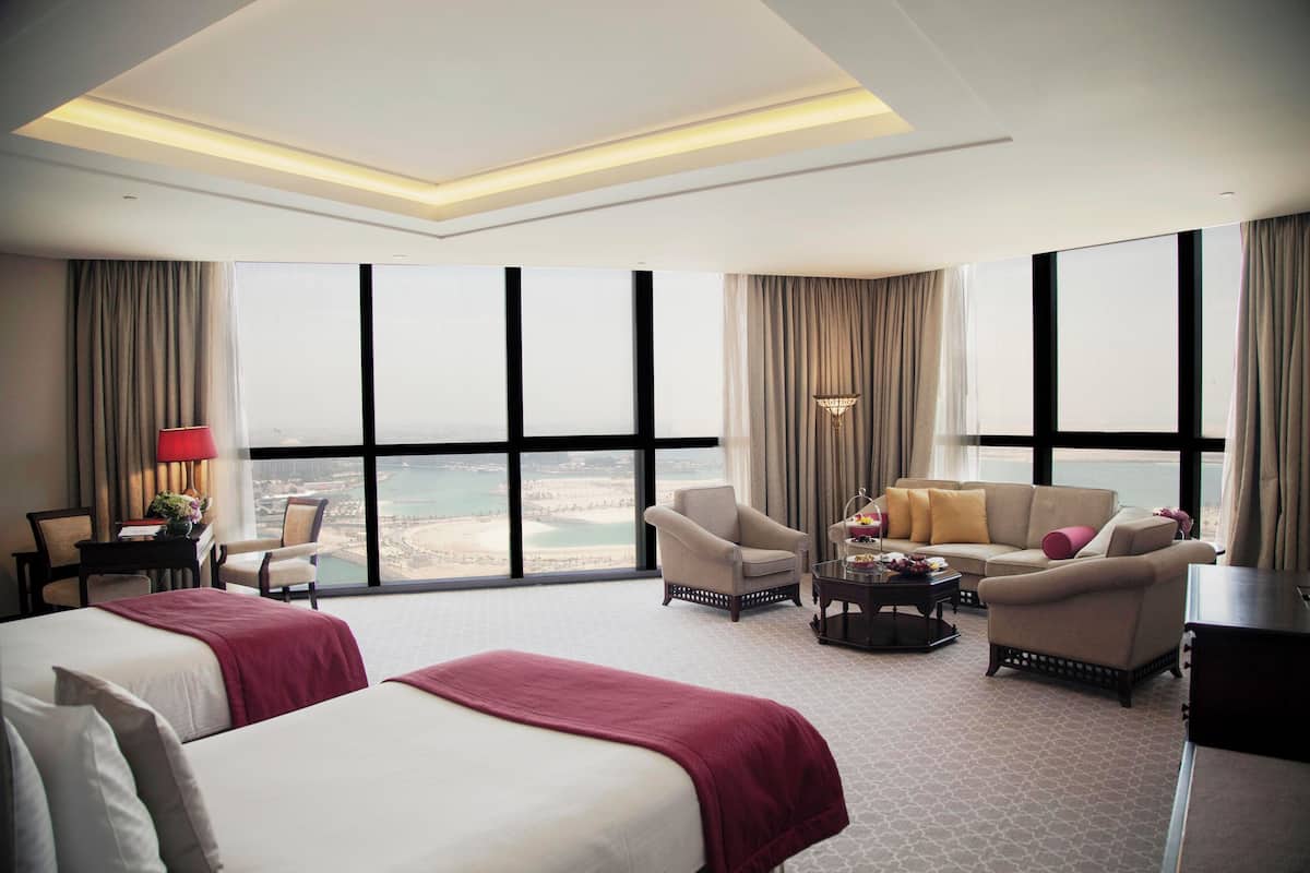 A hotel room with two beds, a sofa, a chair, a desk, and large windows offering a view of the beach.