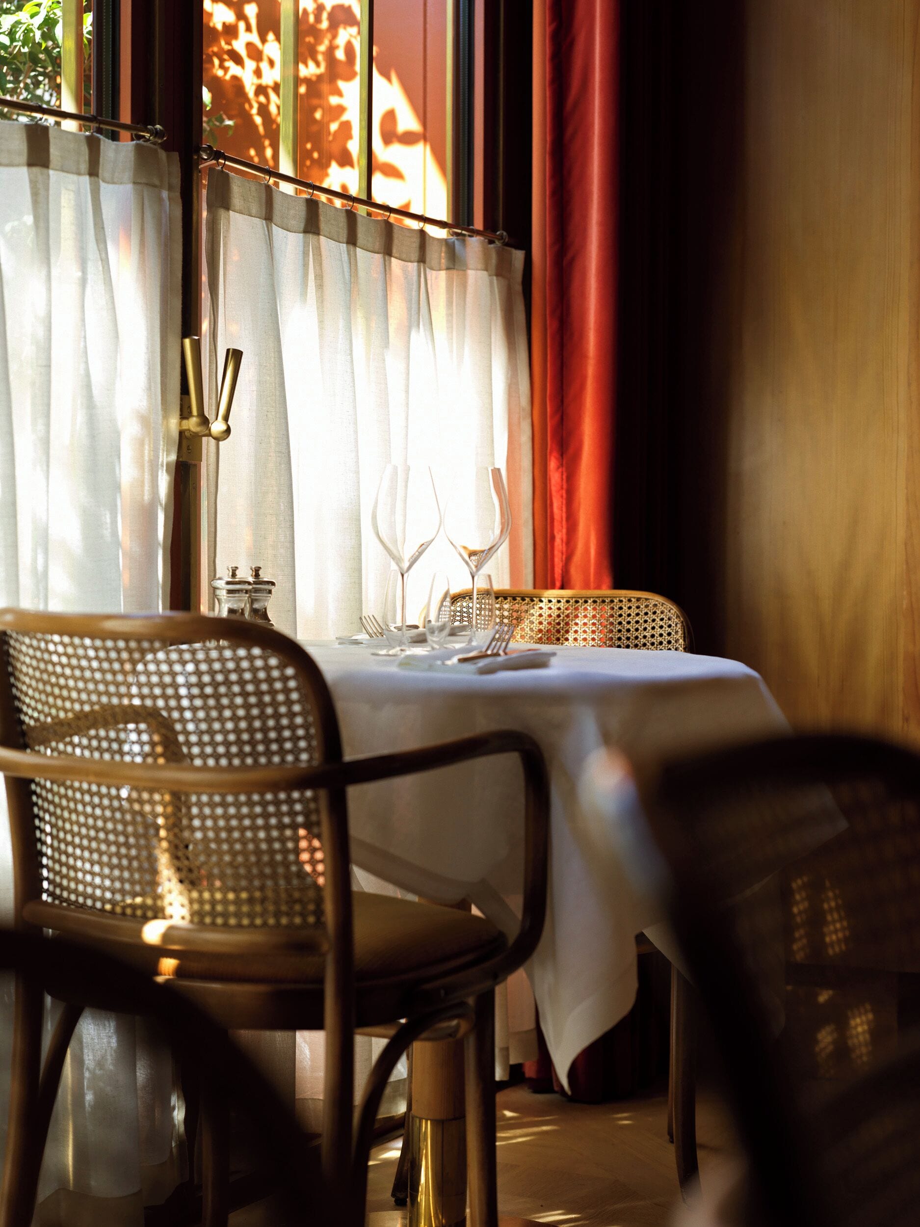 A dining table set with white tablecloth, metal chairs, and a window with sheer curtains and a red drape.
