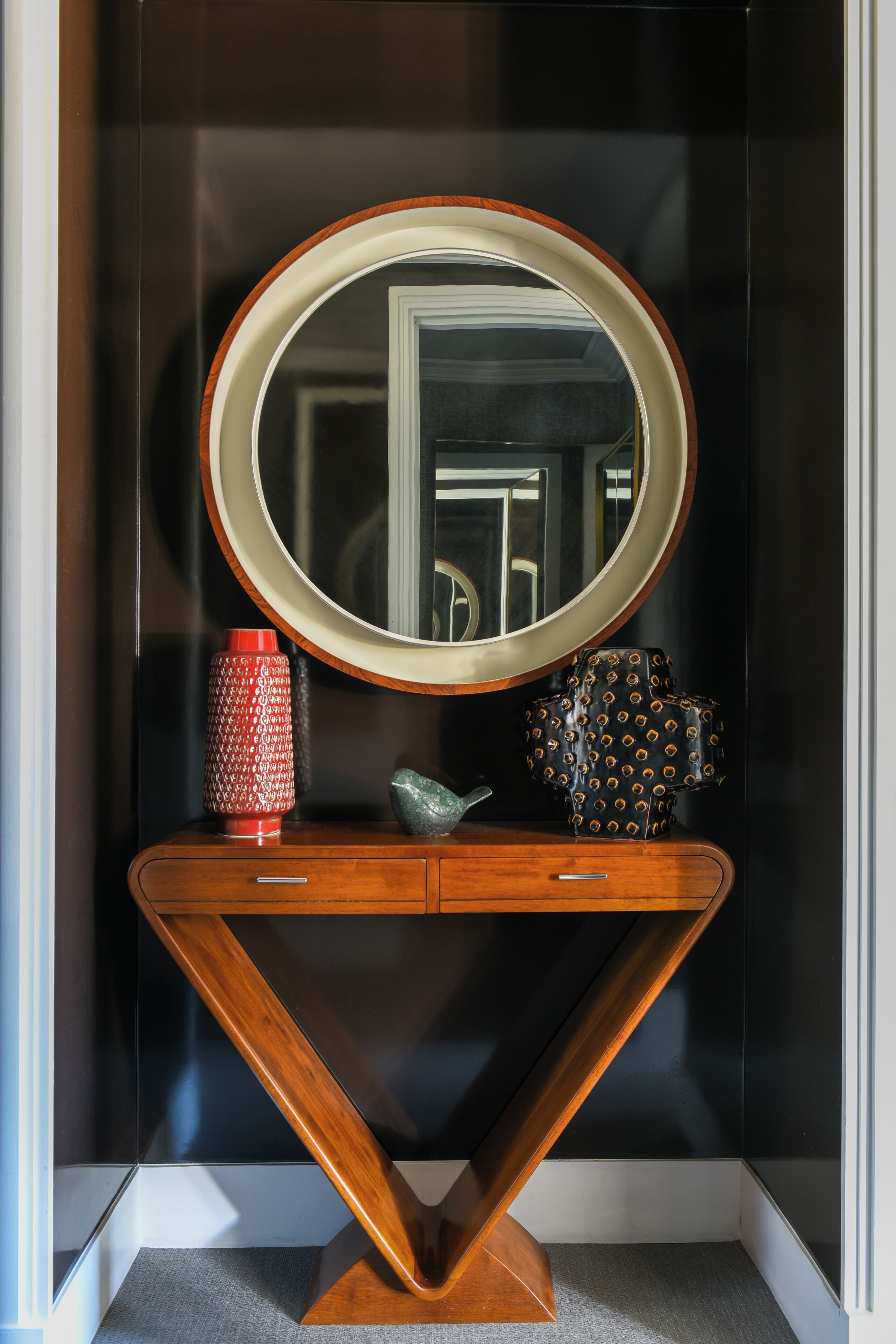 A wooden console table with a mirror, a red vase, and a patterned bowl.