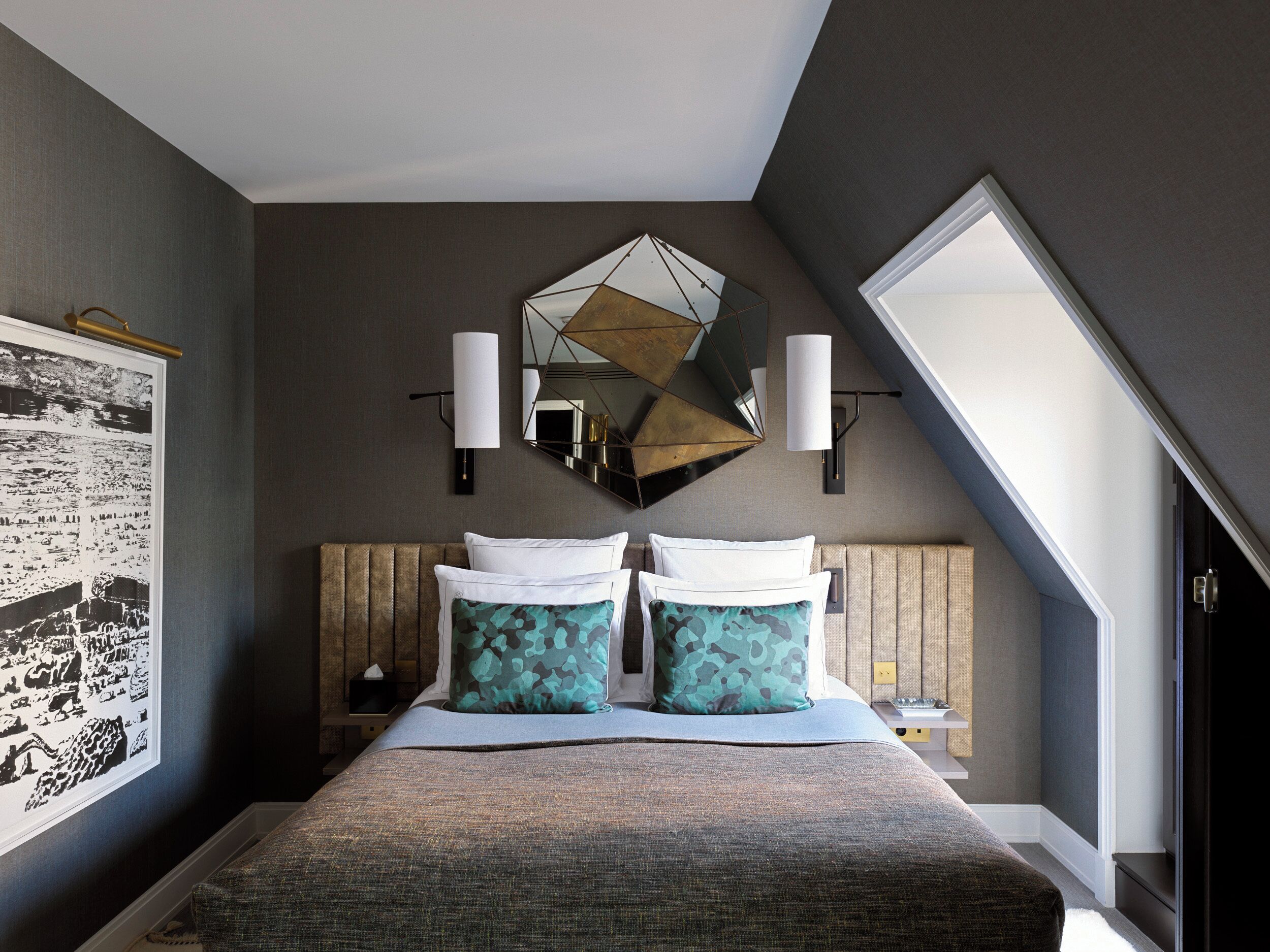 A modern hotel room with a large bed, two wall-mounted lamps, a geometric mirror, and a framed picture on the wall.