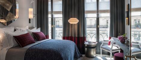 A hotel room with a large bed, a desk with a chair, a small table with a vase, and a view of the city through large windows.