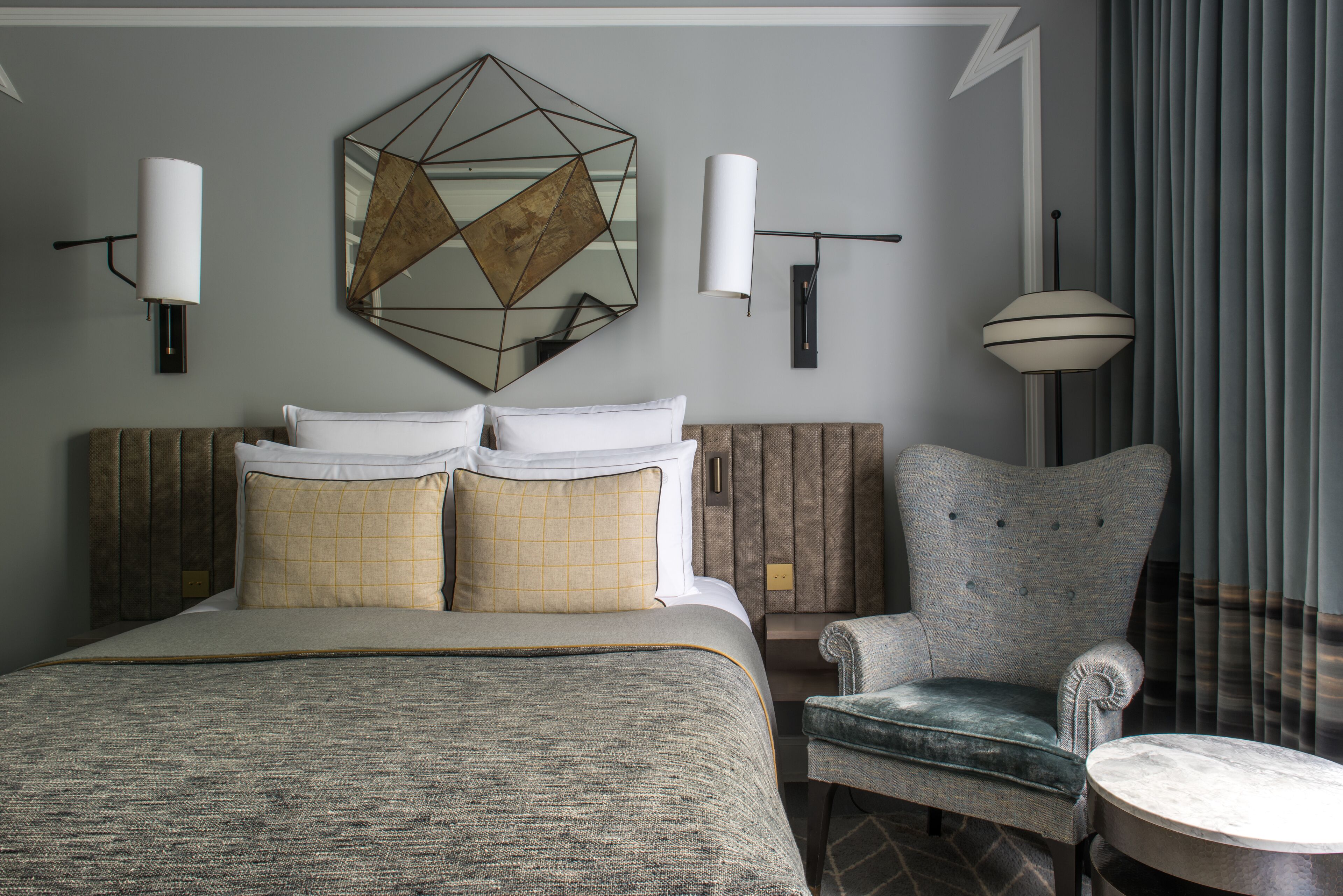 A hotel room with a large bed, a chair, a round table, and a geometric mirror on the wall.