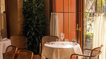 A dining area with a round table, wicker chairs, a potted plant, and a view through a window with curtains.
