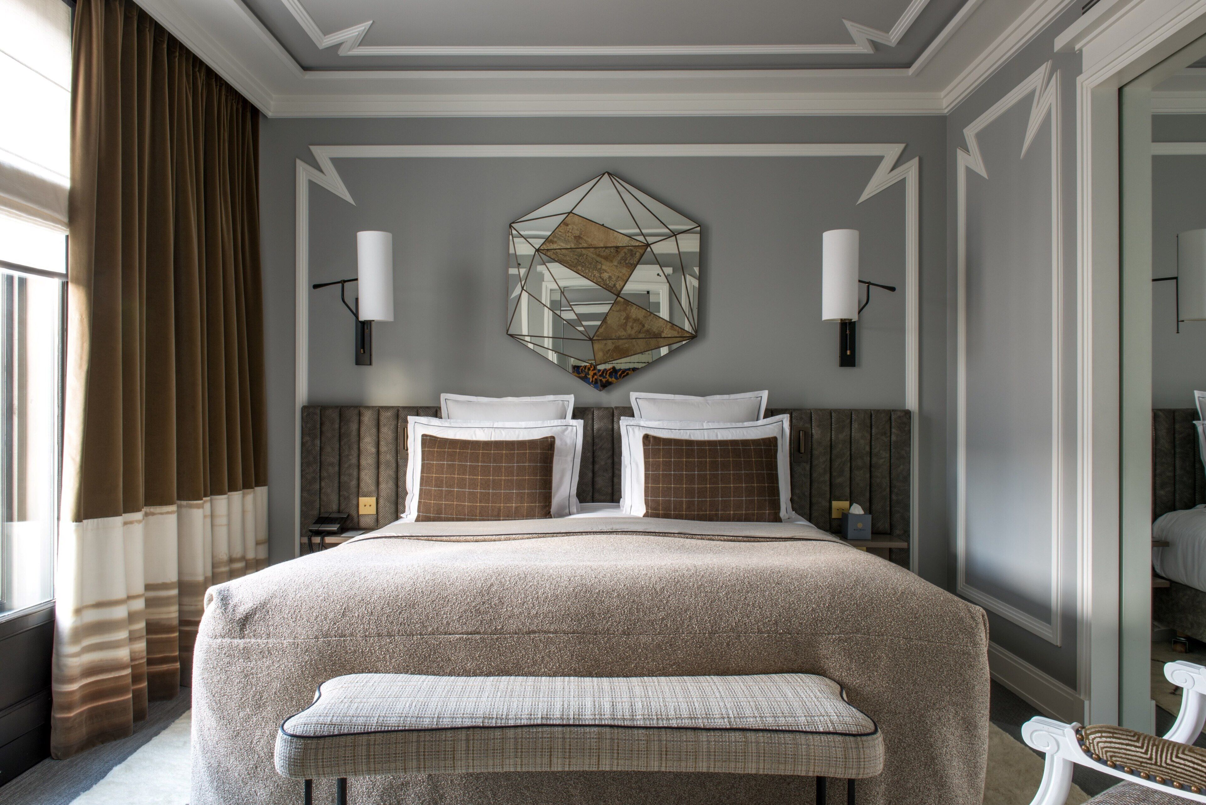 Une chambre d’hôtel avec un grand lit, un banc, un miroir aux motifs géométriques et une fenêtre avec des rideaux.
