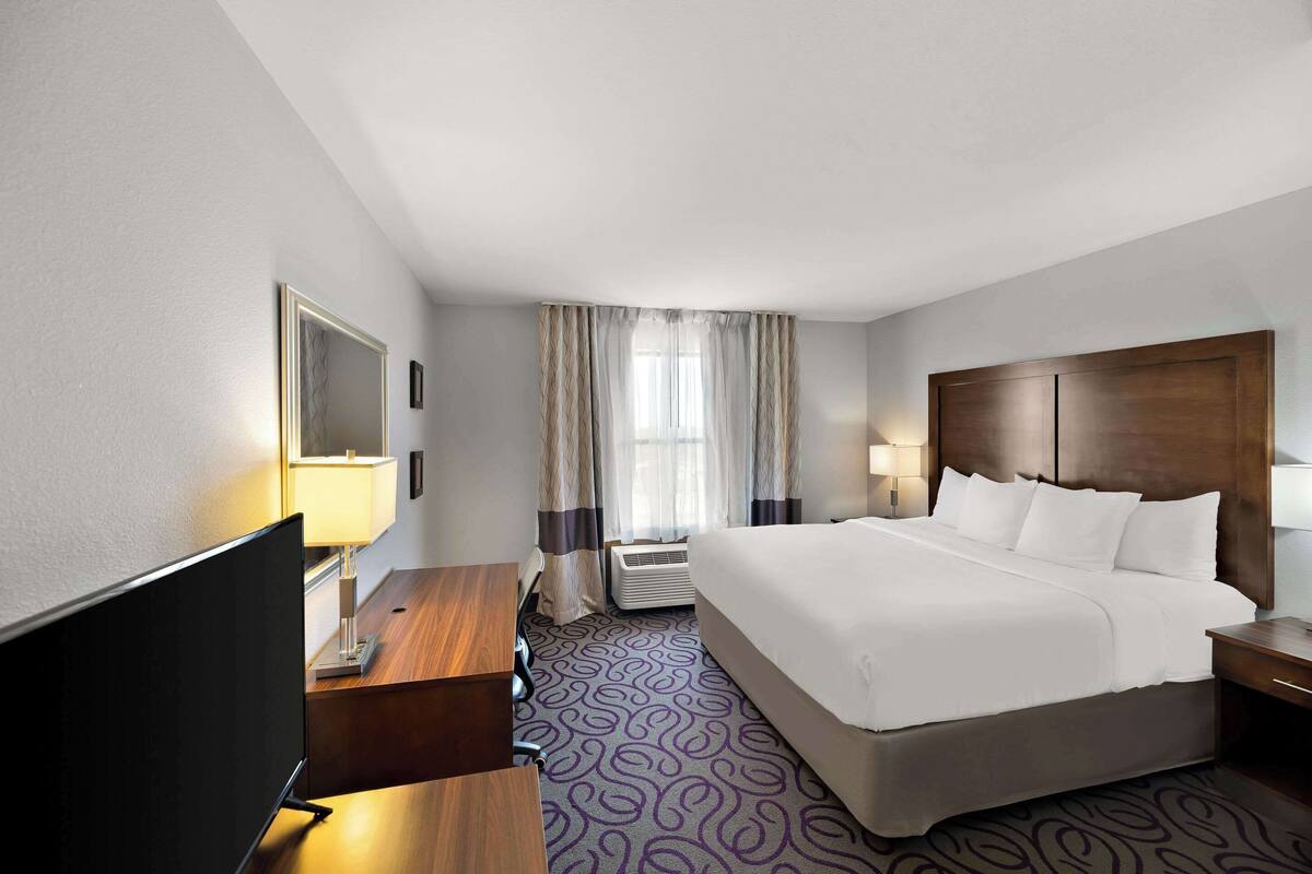 A hotel room with a large bed, a desk with a flat-screen TV, a desk lamp, a window with curtains, and an air conditioning unit.