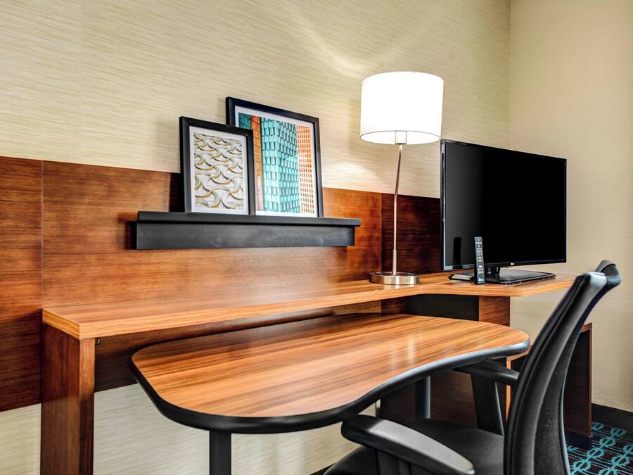 Habitación de hotel con un escritorio de madera, una silla negra, un televisor de pantalla plana, una lámpara y obras de arte enmarcadas en la pared.