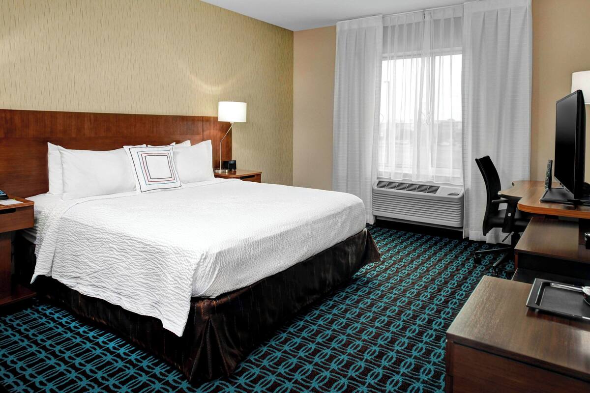A hotel room with a large bed, a desk with a computer, a chair, a TV, and a window with curtains.