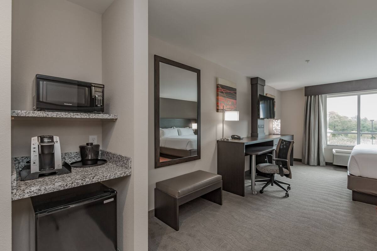 Ein Hotelzimmer mit Küchenzeile, einem Bett, einem Schreibtisch, einem Stuhl und einem großen Fenster.