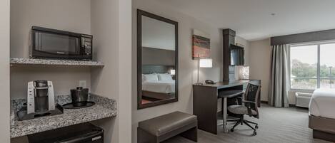 A hotel room with a kitchenette, a bed, a desk, a chair, and a large window.