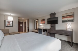 Una habitación de hotel con una cama grande, un escritorio con televisor de pantalla plana, una silla, una lámpara de escritorio y obras de arte en las paredes.