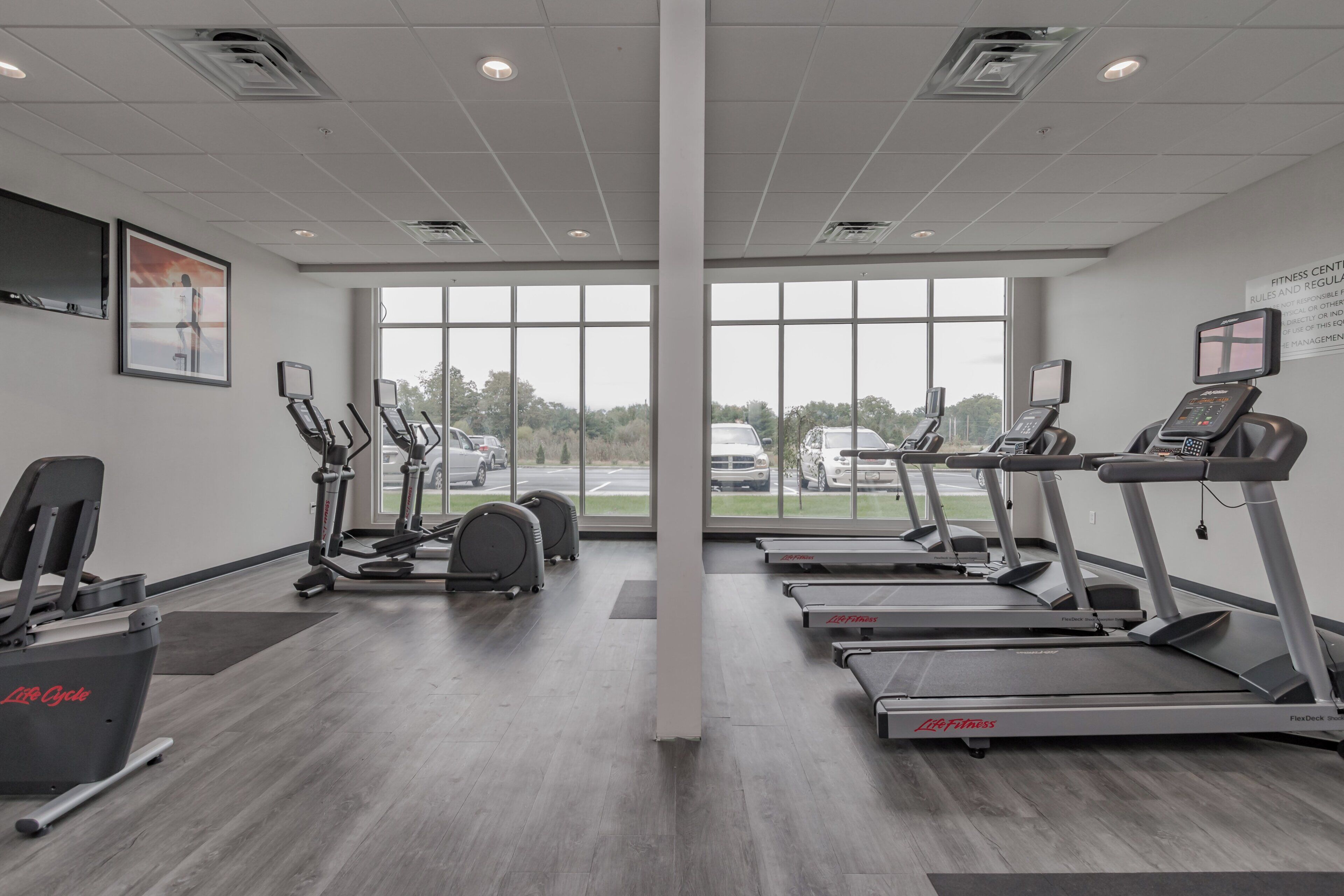 Un gimnasio con cintas de correr, elípticas y una bicicleta estática, ventanas amplias y vista al estacionamiento.