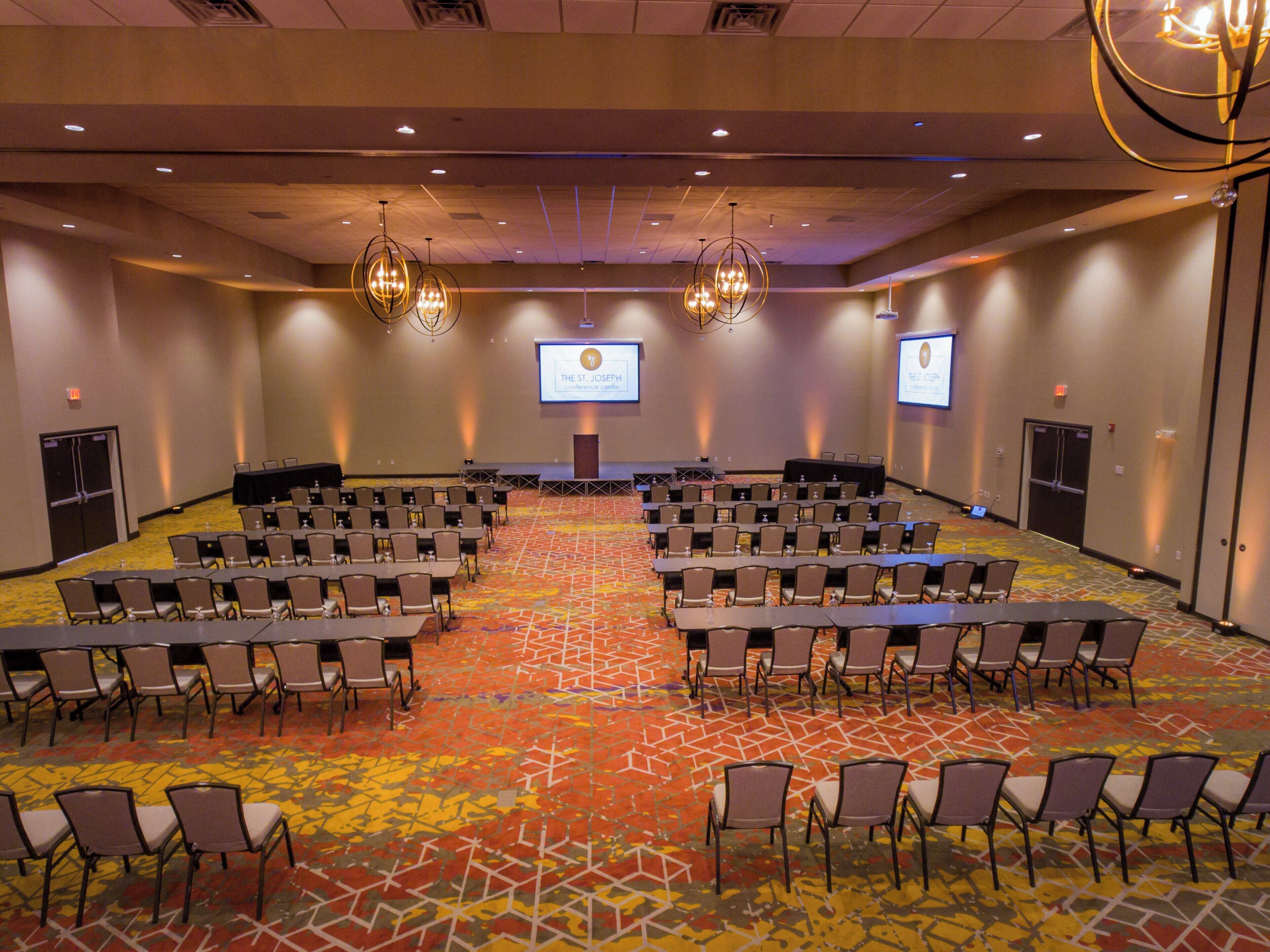 A conference room with rows of tables and chairs, a stage with a screen, and a patterned carpet.