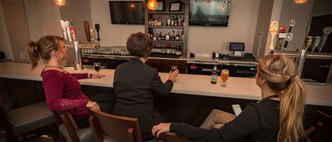 Un coin bar avec trois personnes assises au comptoir, un téléviseur fixé au mur, et diverses bouteilles et verres disposés sur des étagères.