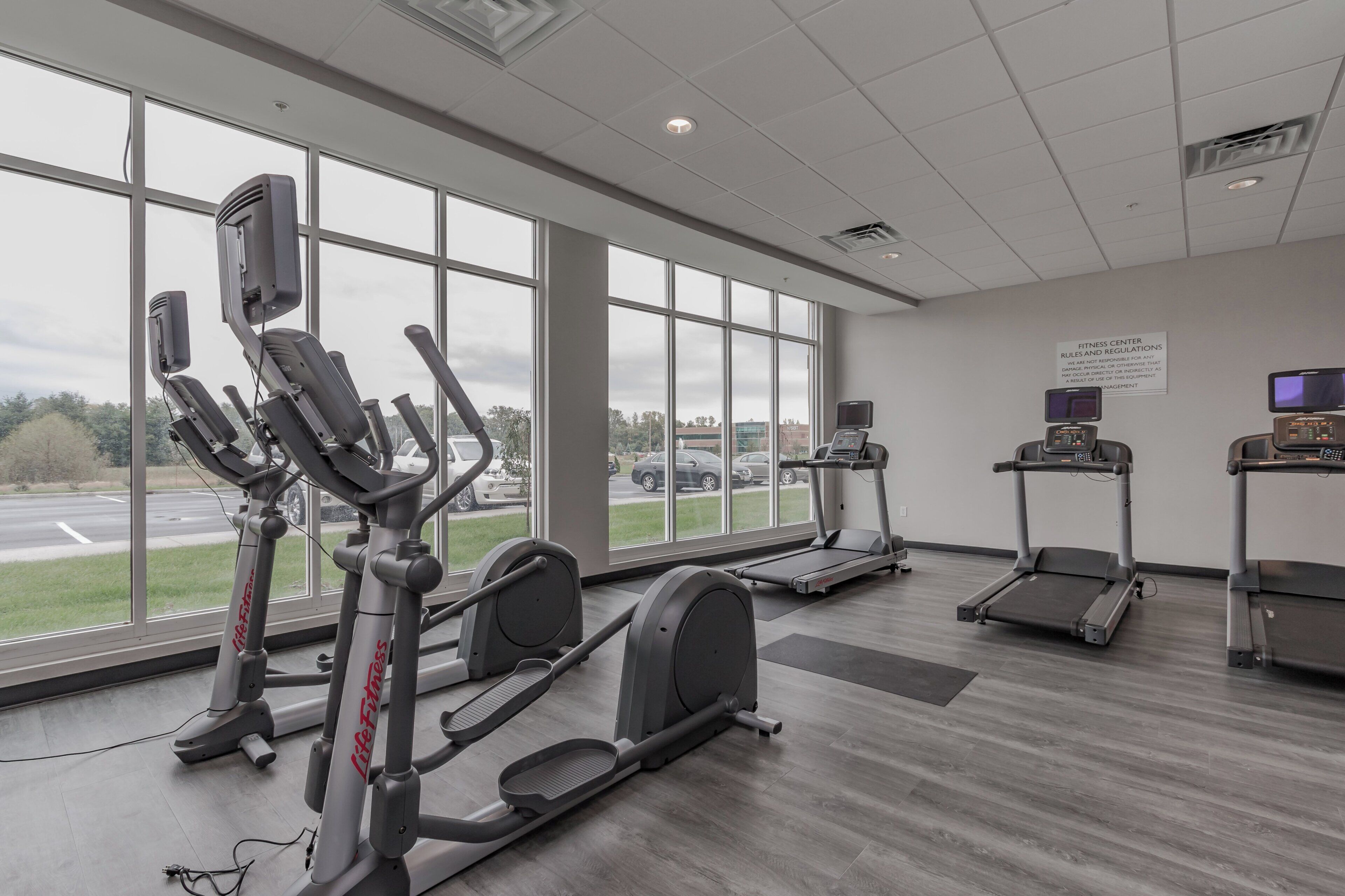 Un gimnasio con equipo cardiovascular, ventanas amplias y un cartel con las normas del centro de acondicionamiento físico.
