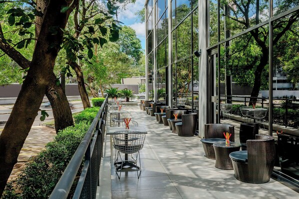 An outdoor seating area with tables and chairs, surrounded by trees and a glass building.