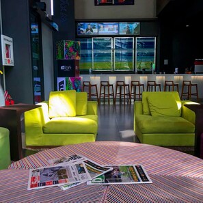 A modern hotel lobby with bright green sofas, a bar area with multiple screens, and a large window offering a view of the outdoors.
