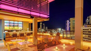 An outdoor bar area with tables and chairs, a bar counter with bottles, and a city skyline view in the background. - Aloft by Marriott Asuncion (Asunción)