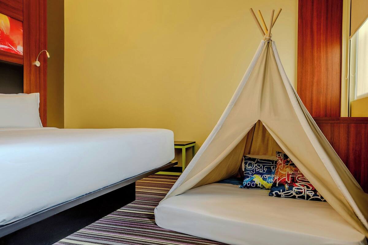 A hotel room with a bed, a striped carpet, a wooden door, and a teepee-style tent with colorful pillows.