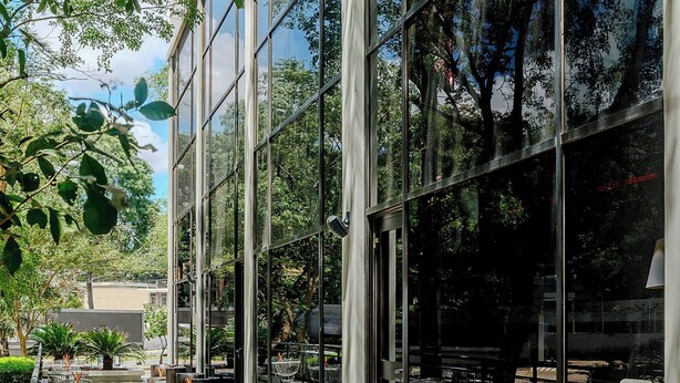 An outdoor cafe with tables and wicker chairs, large glass windows reflecting trees, and a paved walkway. - Aloft by Marriott Asuncion (Asunción)