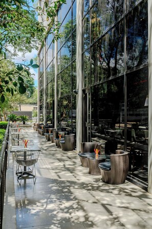 An outdoor cafe with tables and wicker chairs, large glass windows reflecting trees, and a paved walkway.