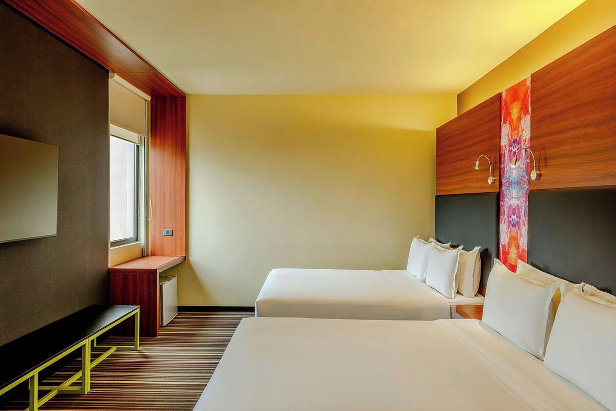 A hotel room with two beds, a desk, a TV, and a window.