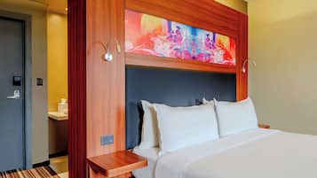 A hotel room with a large bed, a wooden headboard, a colorful abstract painting above the bed, and a small bedside table.