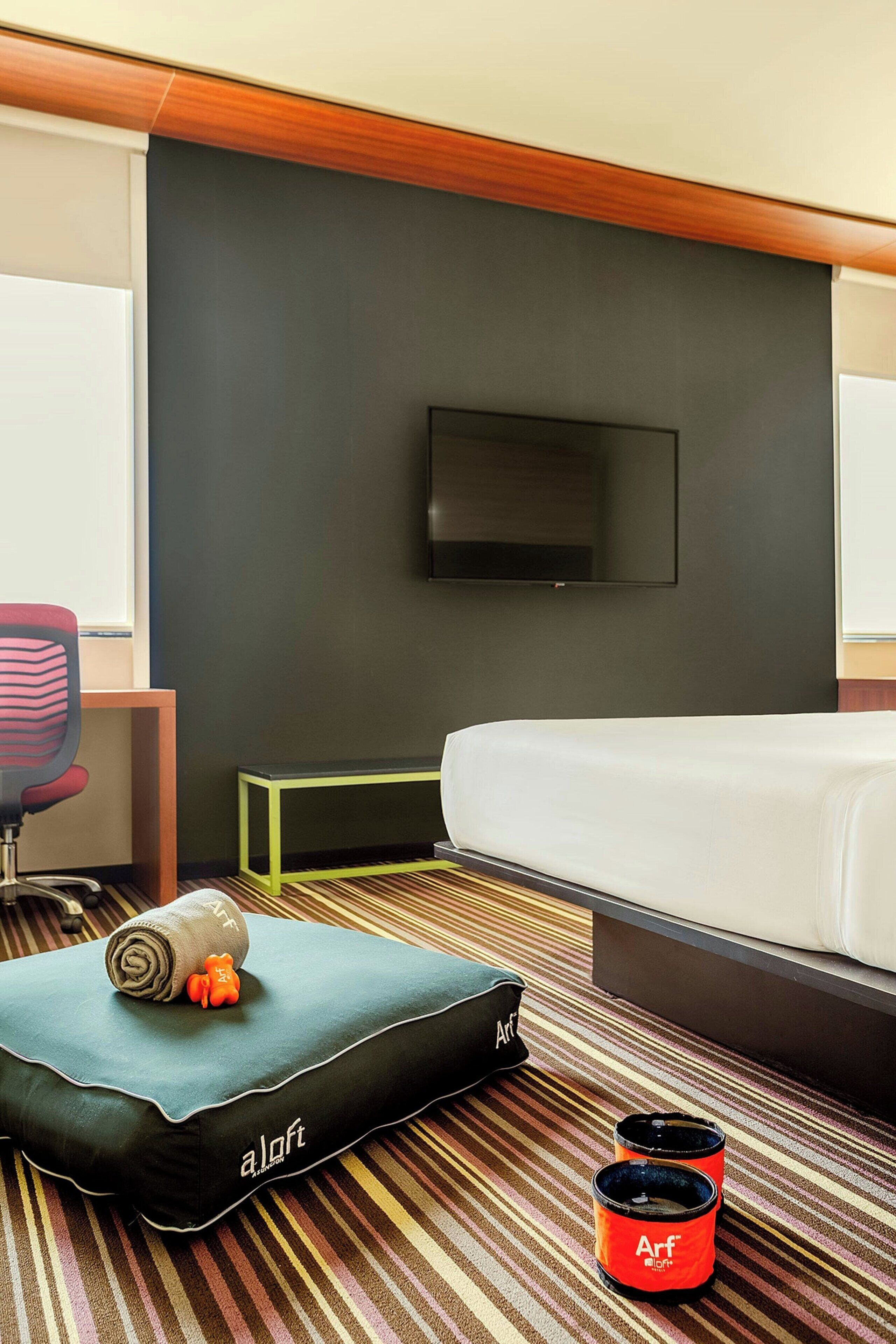 A hotel room with a bed, a TV mounted on the wall, a striped carpet, a small table, and a pillow with a rolled towel and a stuffed toy.