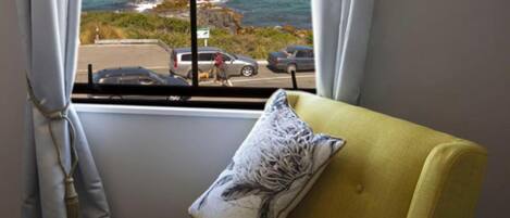 A yellow armchair with a patterned pillow, positioned next to a window with a view of the sea and a parking area.