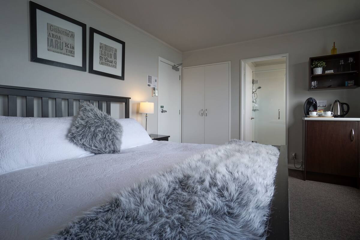 A neatly arranged bedroom with a bed, a fluffy grey throw, framed pictures on the wall, a bedside table with a lamp, and a wardrobe.